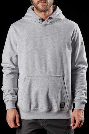 A man is standing against a black background wearing a light grey FXD WF-1 Hoodie, featuring a front pocket. The sweatshirt, crafted from windproof fleece, has a small green tag with the letters "FXD" on the pocket and subtle branding on the left sleeve. The man has a beard and short hair.