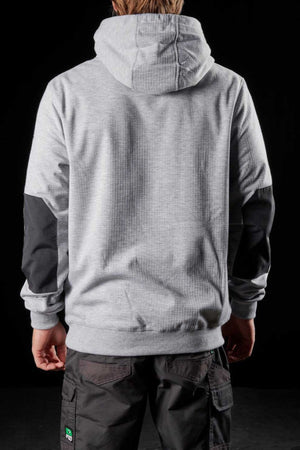 A person stands with their back to the camera, donning an FXD WF-1 Hoodie in light gray, featuring a hood up and black patches on the sleeves. They are also wearing dark-colored pants adorned with a green logo near the pocket. The background is completely black.