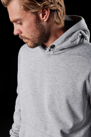 A person with blond hair and a beard is looking downwards while wearing a light gray FXD WF-1 Hoodie. The black background accentuates the subject's profile and the texture of the windproof fleece hoodie.