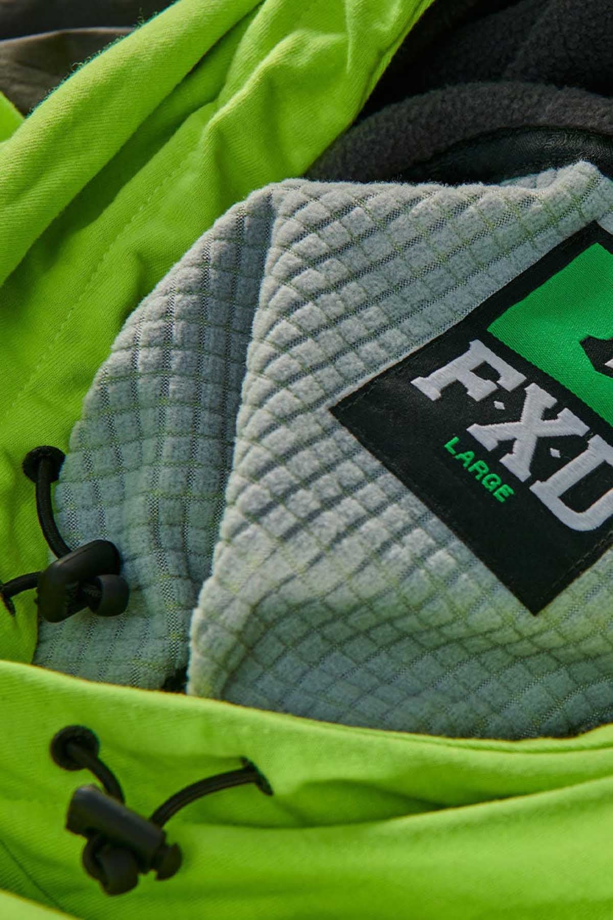Close-up of a heather grey garment label on a textured fabric with the word "LARGE" and "FXD" printed on it. The background features lime green fabric with black drawstrings, partially covering the FXD WF-1 Hoodie which is water resistant.