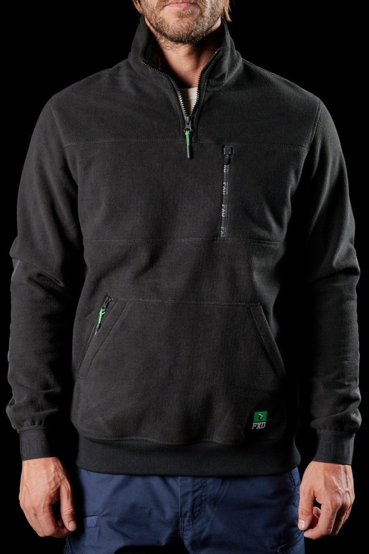 A man with light facial hair stands against a black background wearing the FXD WF-2 Fleece, a black, windproof pullover featuring a quarter-zip, chest pocket, and front pouch pocket.
