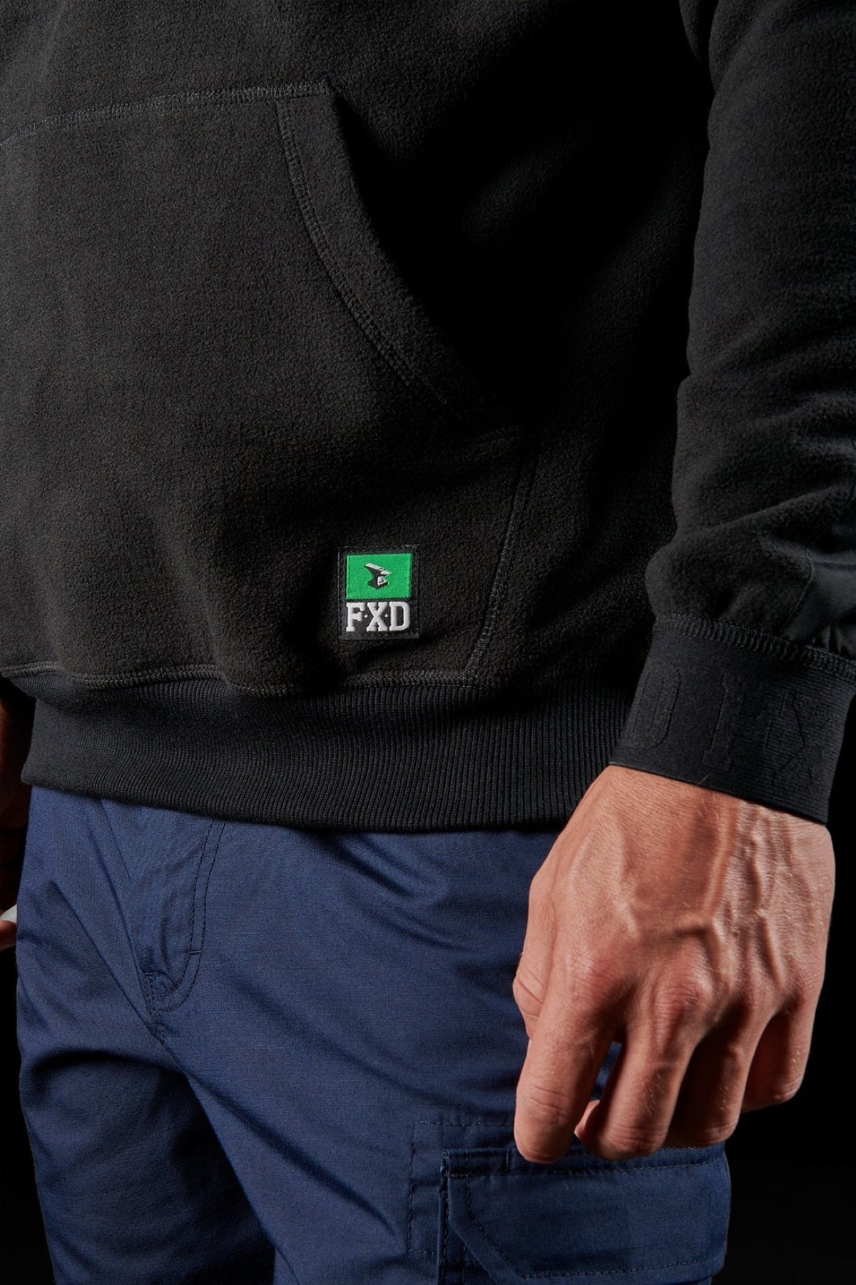 A person wearing a black FXD WF-2 Fleece hoodie with windproof fleece and blue cargo pants, shown from the waist down with one hand visible. The FXD logo appears on the hoodie's pocket.