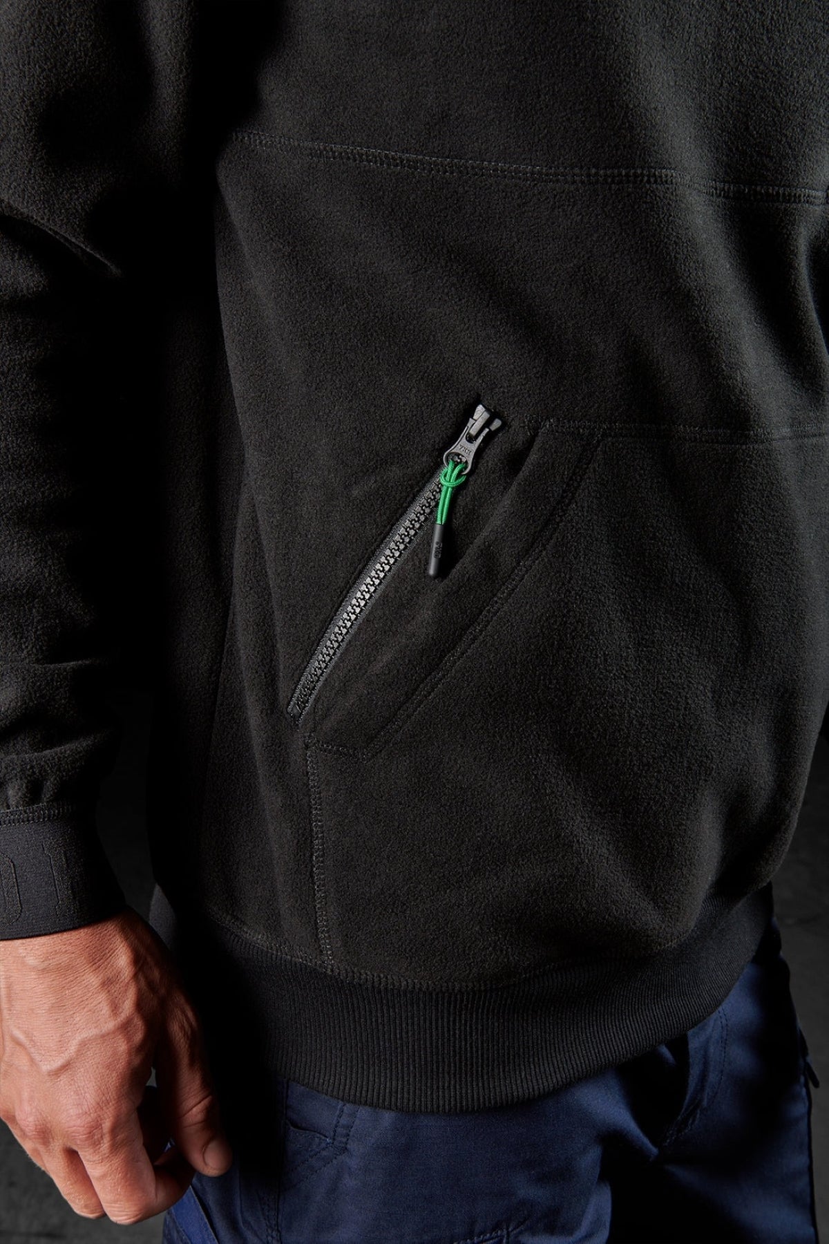 A close-up of a person wearing the FXD WF-2 Fleece, a black water-resistant, windproof jacket with a zippered pocket, paired with dark blue pants. Their left hand is relaxed at their side.