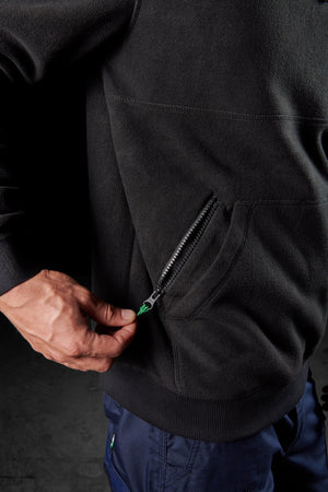 A person’s hand pulls the zipper of a pocket on a black FXD WF-2 Fleece jacket, which is water resistant and windproof. The person is wearing blue pants, and the dark background is out of focus.