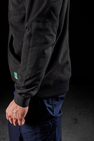 A person wearing the FXD WF-2 Fleece, a black windproof sweater with reinforced elbow patches, and dark blue pants stands against a dark background with their left arm relaxed at their side.