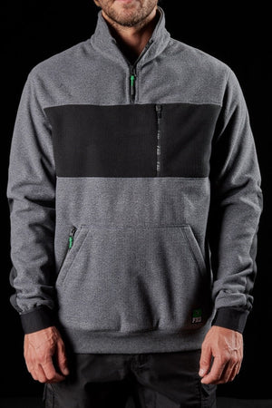 A man stands against a black background wearing the FXD WF-2 Fleece, a gray pullover with a black chest panel, zippered collar, and front pocket. This windproof fleece keeps him comfortable, his face partially visible and hands relaxed.
