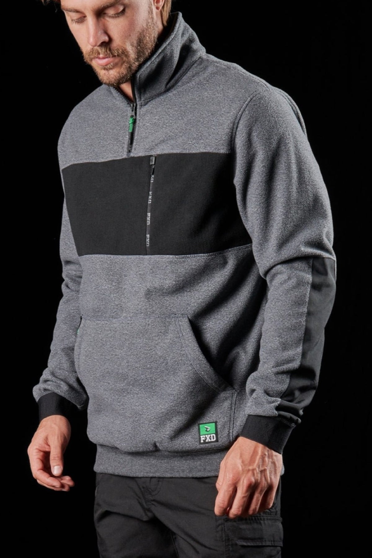 A man wears an FXD WF-2 Fleece—a grey pullover with black chest panel, green logo patch, and water-resistant windproof fleece—standing against a black background, gazing down with one hand by his side and one in the pouch pocket.