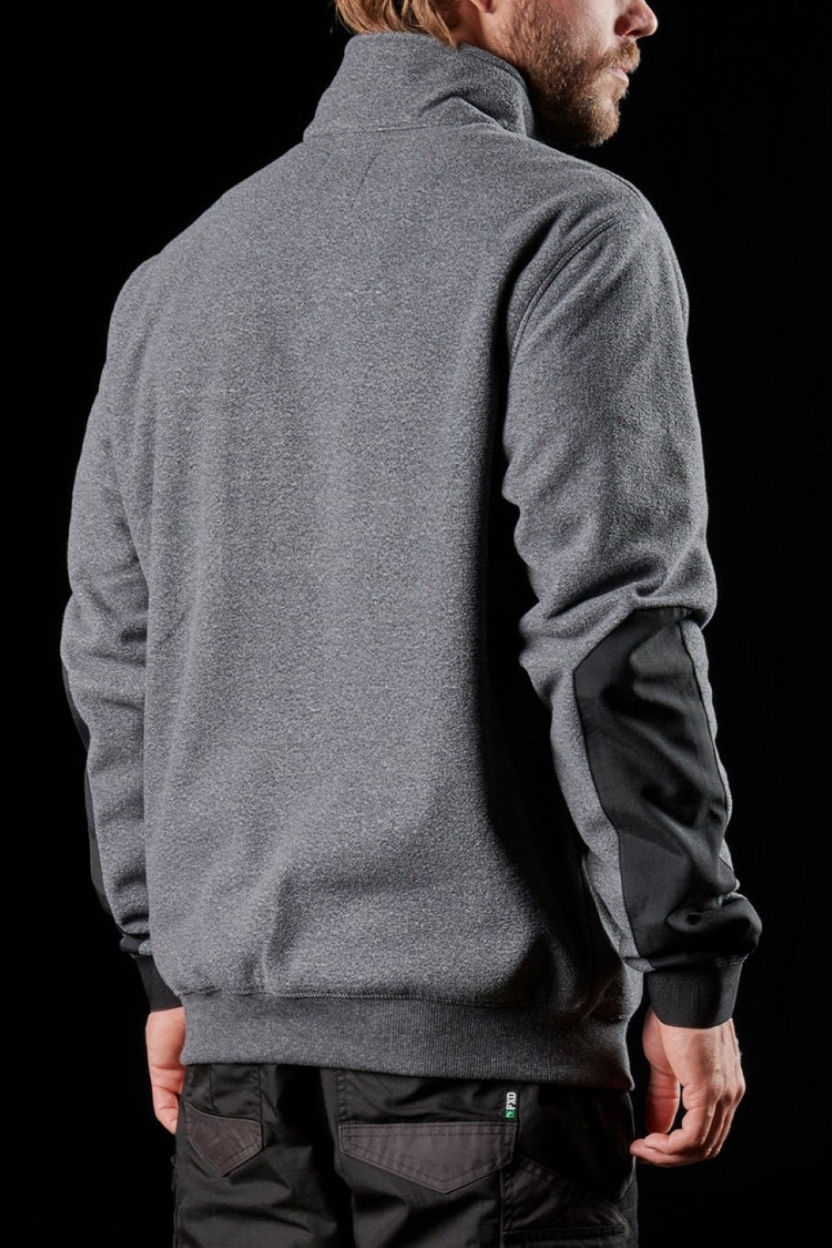A man with light hair and a beard faces away, wearing the FXD WF-2 Fleece jacket in gray with black elbow patches and black pants, set against a black background.