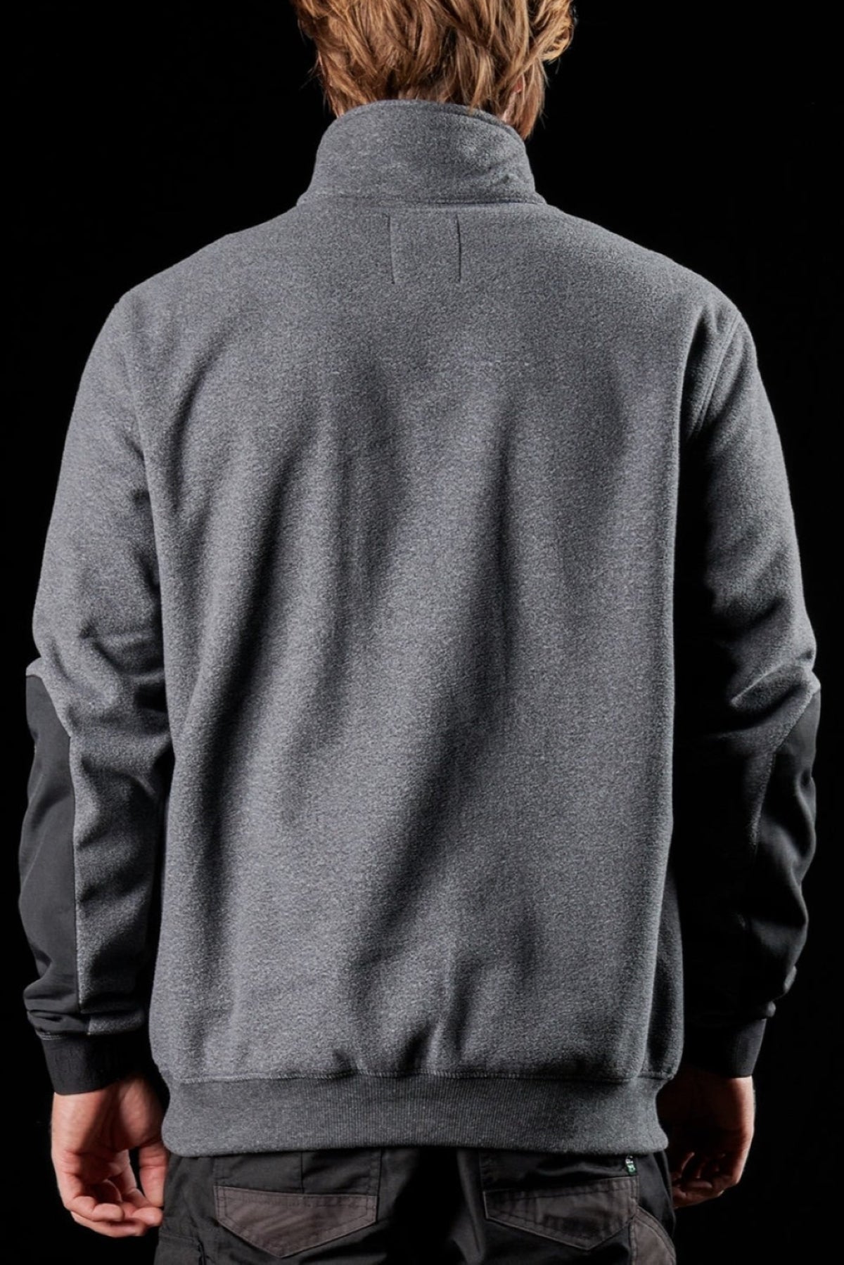 A person with light brown hair is seen from behind wearing the FXD WF-2 Fleece, a gray windproof jacket with darker fabric on the elbows, set against a solid black background.