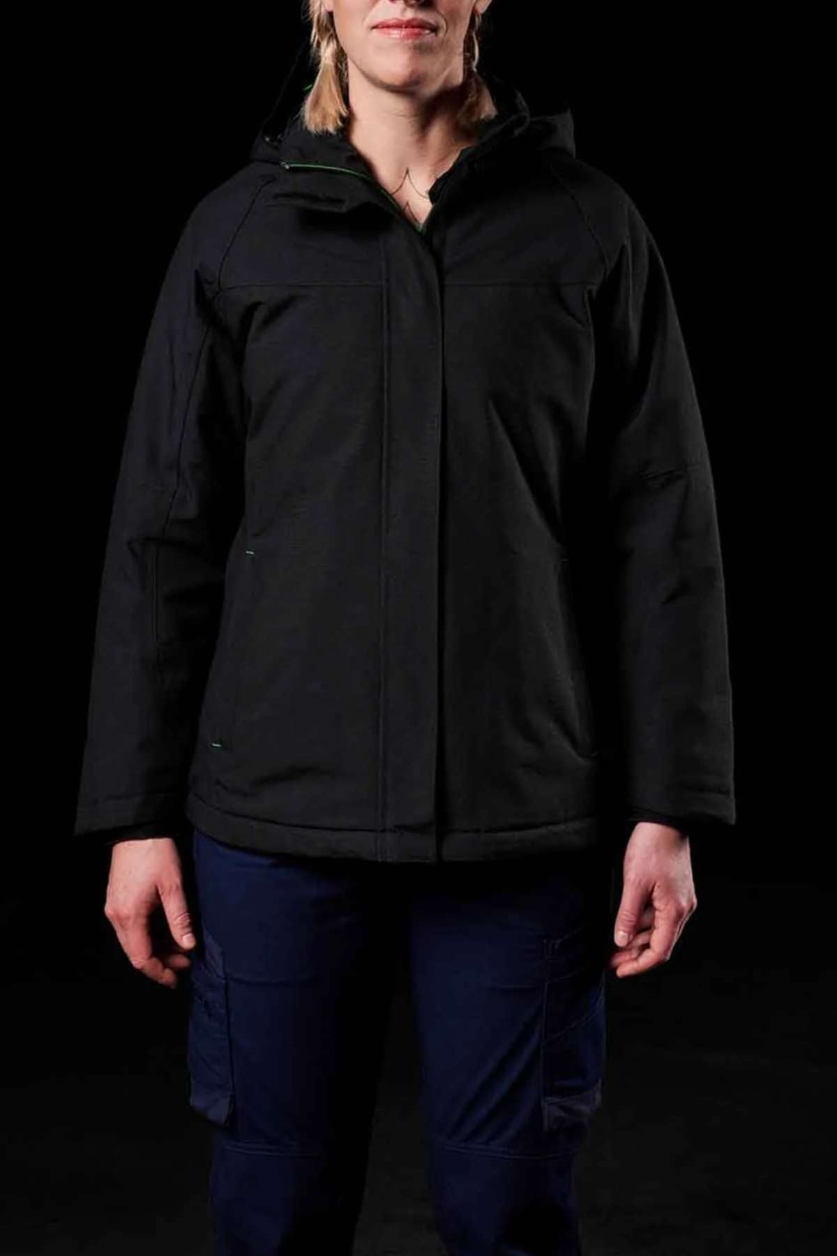 A person wears the FXD WO-1W Womens Insulated Work Jacket, black in color, with dark blue pants against a black background. Their head is partly out of frame and their hands are relaxed by their sides.