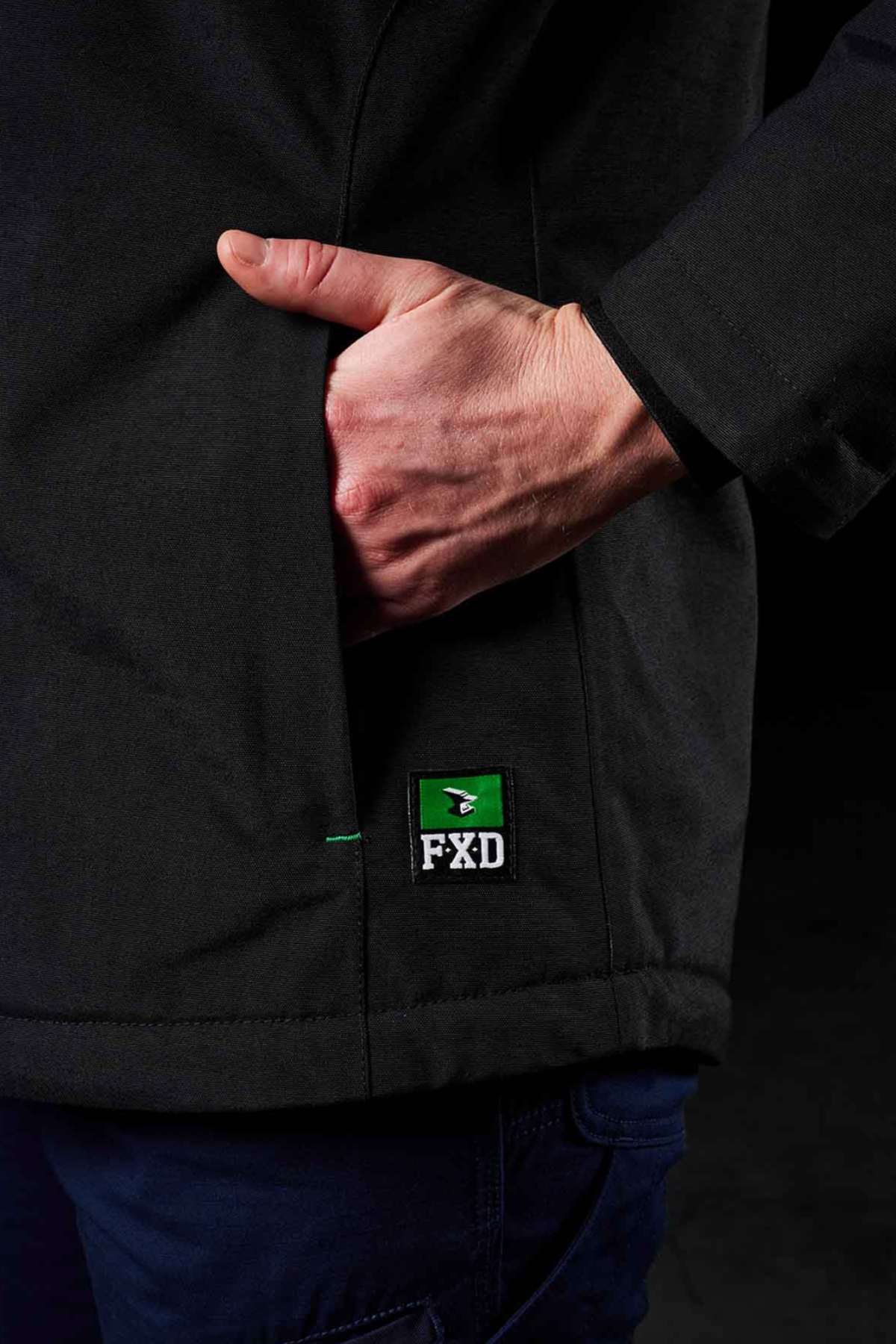 A person wears the FXD WO-1W Womens Insulated Work Jacket—black, nylon, with right hand in pocket. A green and white FXD logo patch appears near the pocket. The background is dark.