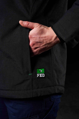 A person wears the FXD WO-1W Womens Insulated Work Jacket—black, nylon, with right hand in pocket. A green and white FXD logo patch appears near the pocket. The background is dark.