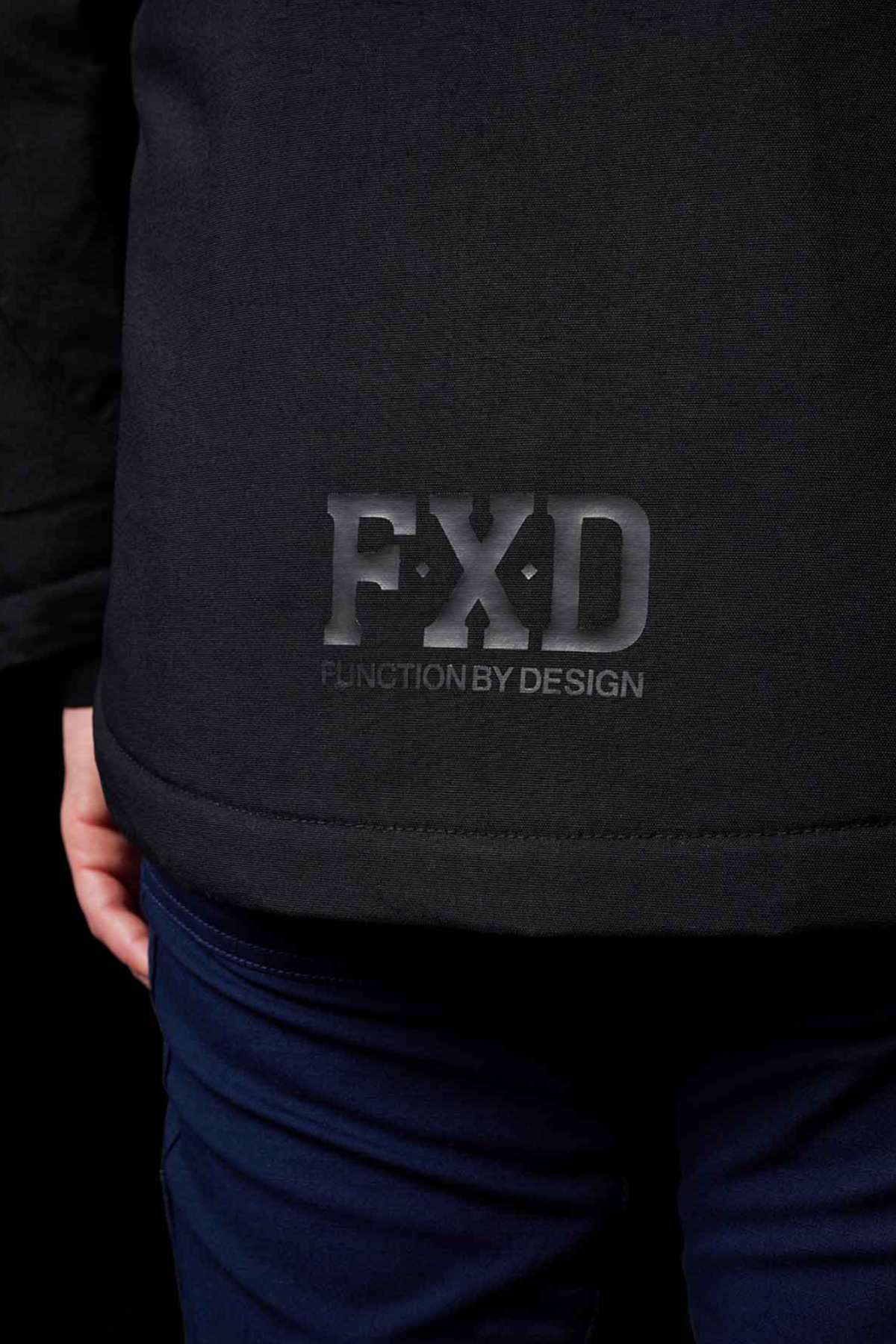 A close-up of someone wearing the FXD WO-1W Womens Insulated Work Jacket with "FXD FUNCTION BY DESIGN" on the lower back. Their hand and the top of dark blue pants are partially visible.