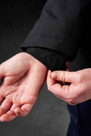 A close-up shows a person adjusting the cuff of the FXD WO-1W Womens Insulated Work Jacket with one hand, while the other hand is open, revealing the wrist and forearm against a dark background.