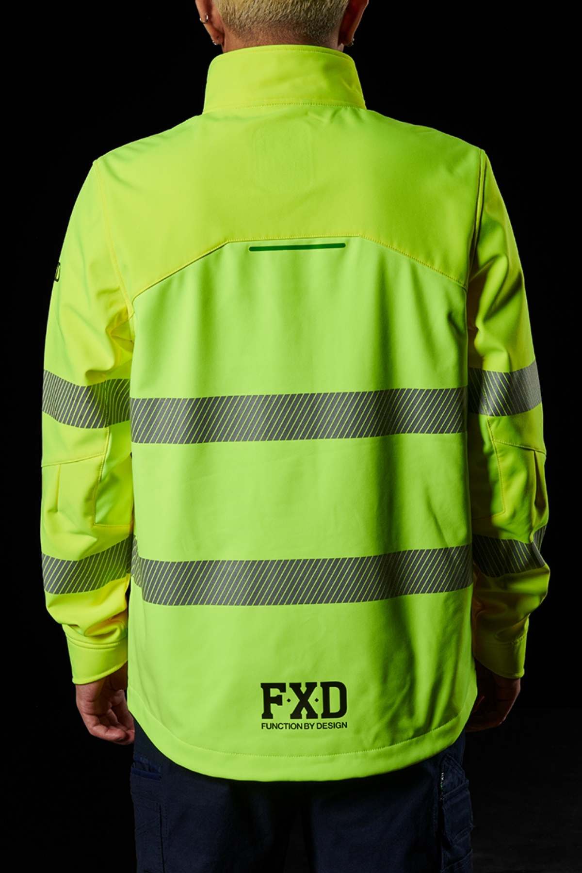 Against a black background, a person wears the FXD WO-3T Hi Vis Soft Shell Work Jacket in neon yellow with silver reflective stripes. The wind and water-resistant design features bold "FXD" lettering at the bottom, ensuring high visibility and reliability.