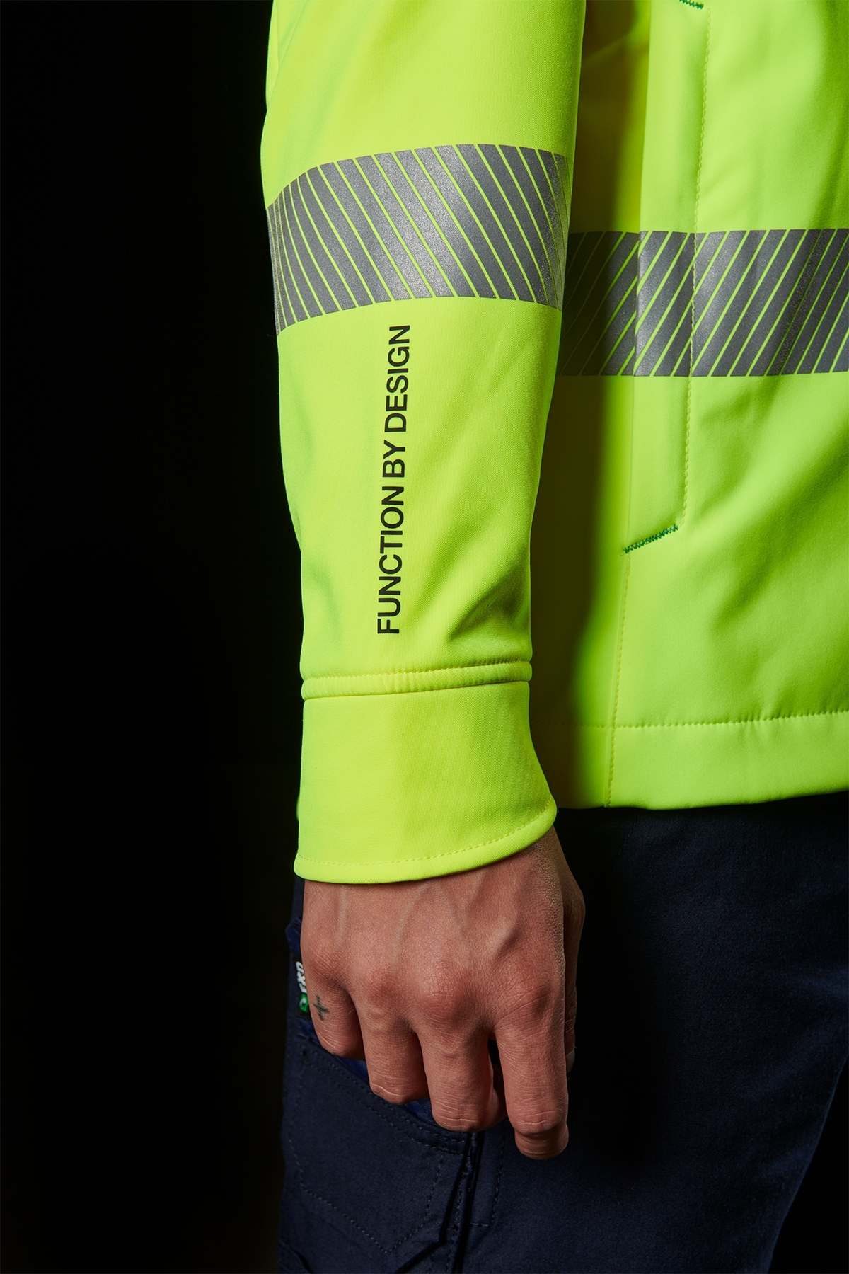 Against a solid black background, a person confidently wears the FXD WO-3T Hi Vis Soft Shell Work Jacket in neon yellow with reflective stripes. The sleeve bears the text "FUNCTION BY DESIGN," showcasing its wind and water-resistant features as their hand rests by their side.