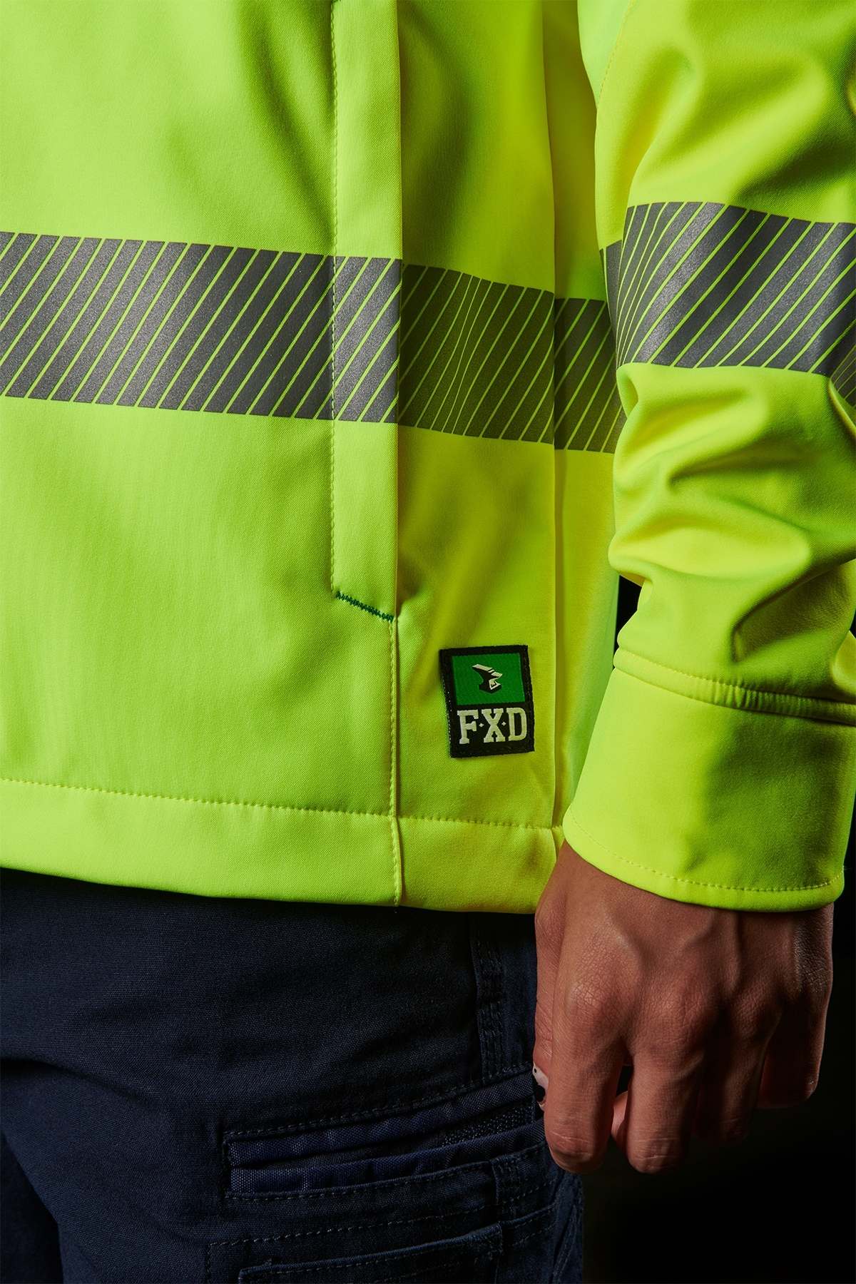 A person wears the FXD WO-3T Hi Vis Soft Shell Work Jacket in bright yellow with reflective stripes for wind and water resistance. Their right hand rests by their side against a dark background, showcasing the "FXD" label.