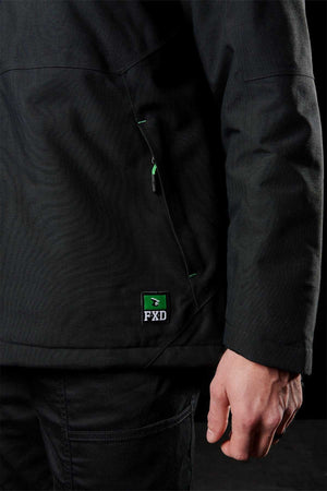 A person wearing a black FXD WO-1 Insulated Work Jacket and black pants stands with their right hand by their side. The FXD logo patch is visible on the lower left of the jacket, near a zippered pocket.