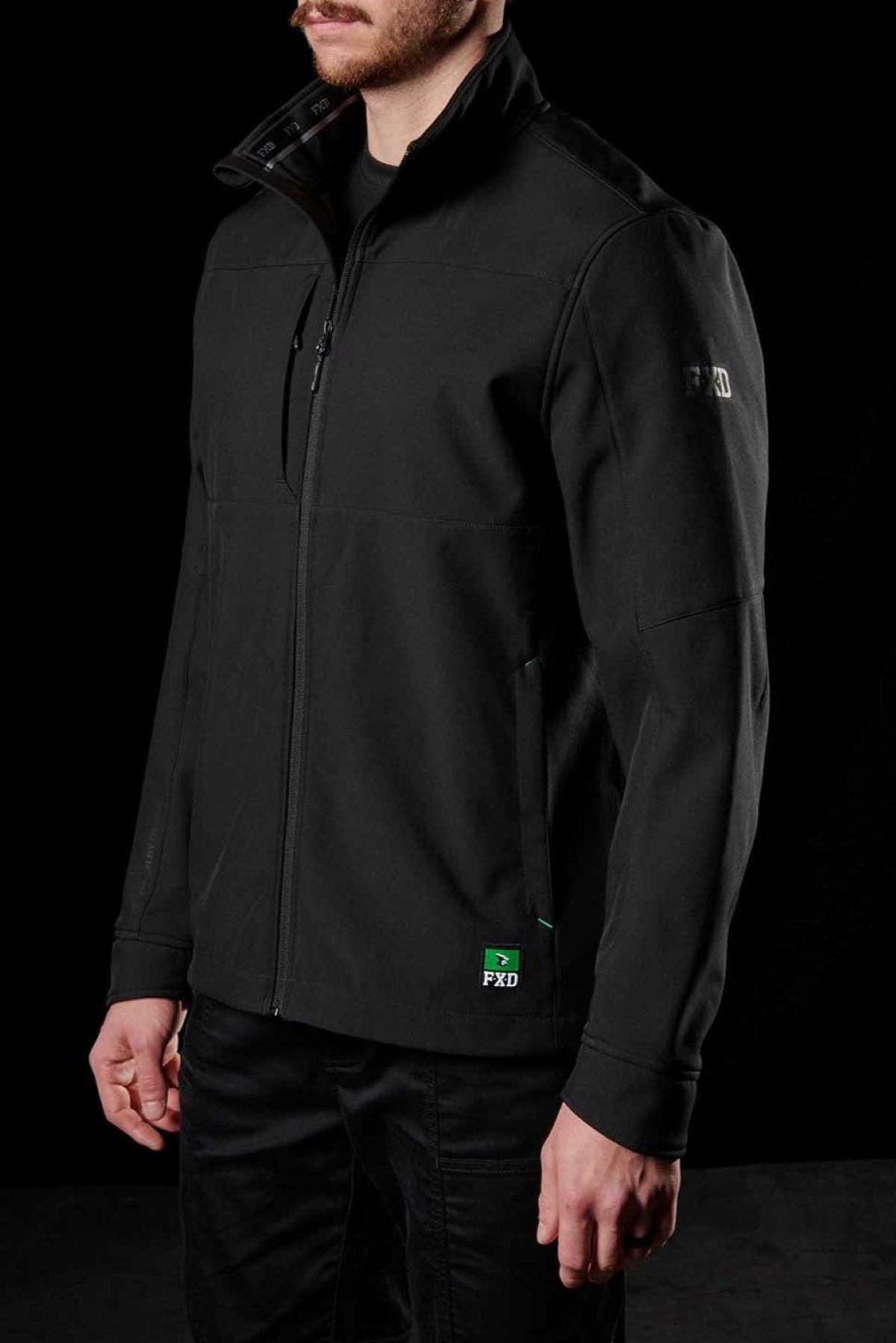 A man showcases the FXD WO-3 Soft Shell Work Jacket, featuring a high collar and subtle green chest logo. Made from breathable, wind and water-resistant fabric with 50+ UPF protection, its sleek design is highlighted against a completely black background.