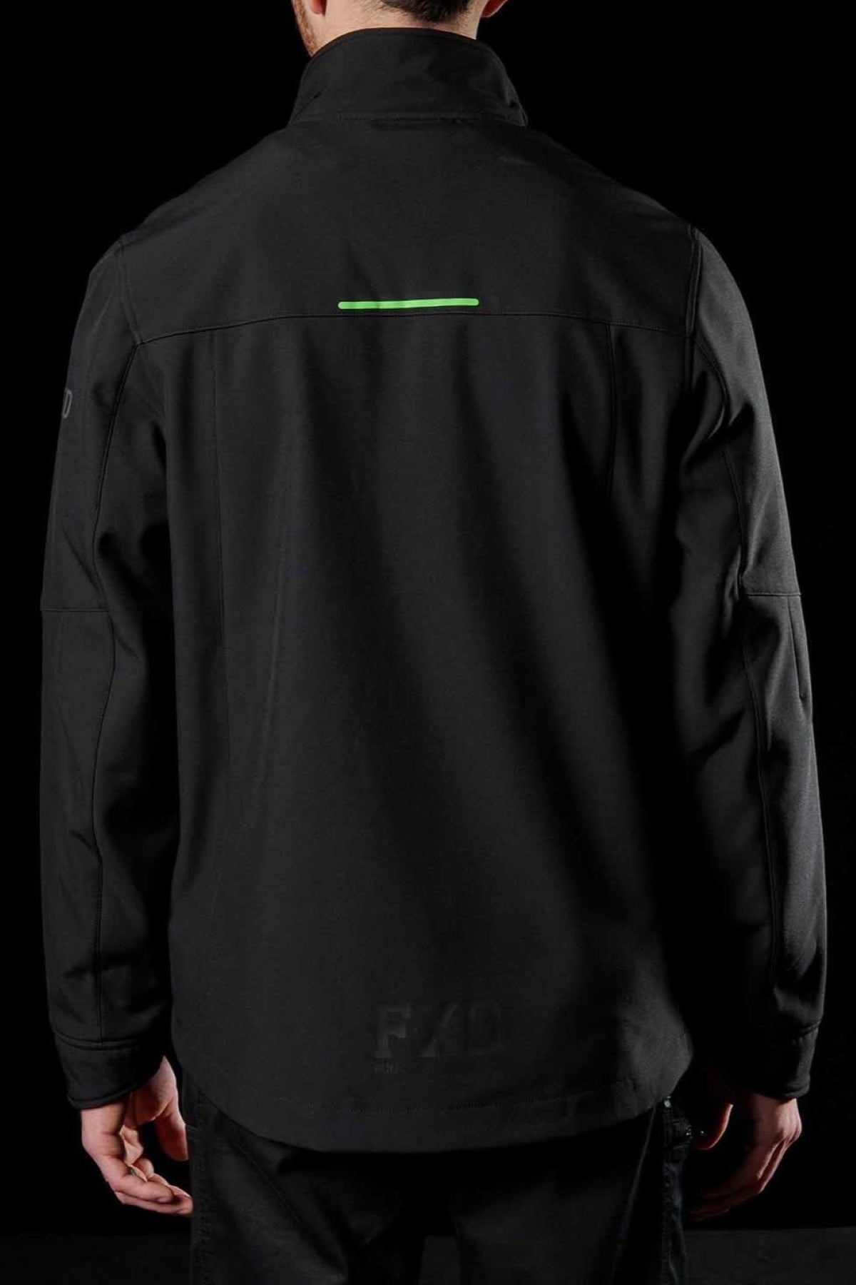 A person wears the FXD WO-3 Soft Shell Work Jacket, featuring a green horizontal stripe on the upper back. This breathable wind and water-resistant jacket offers 50+ UPF protection, visible seams, and a subtle "FXD" print near the bottom, standing out against its solid black background.