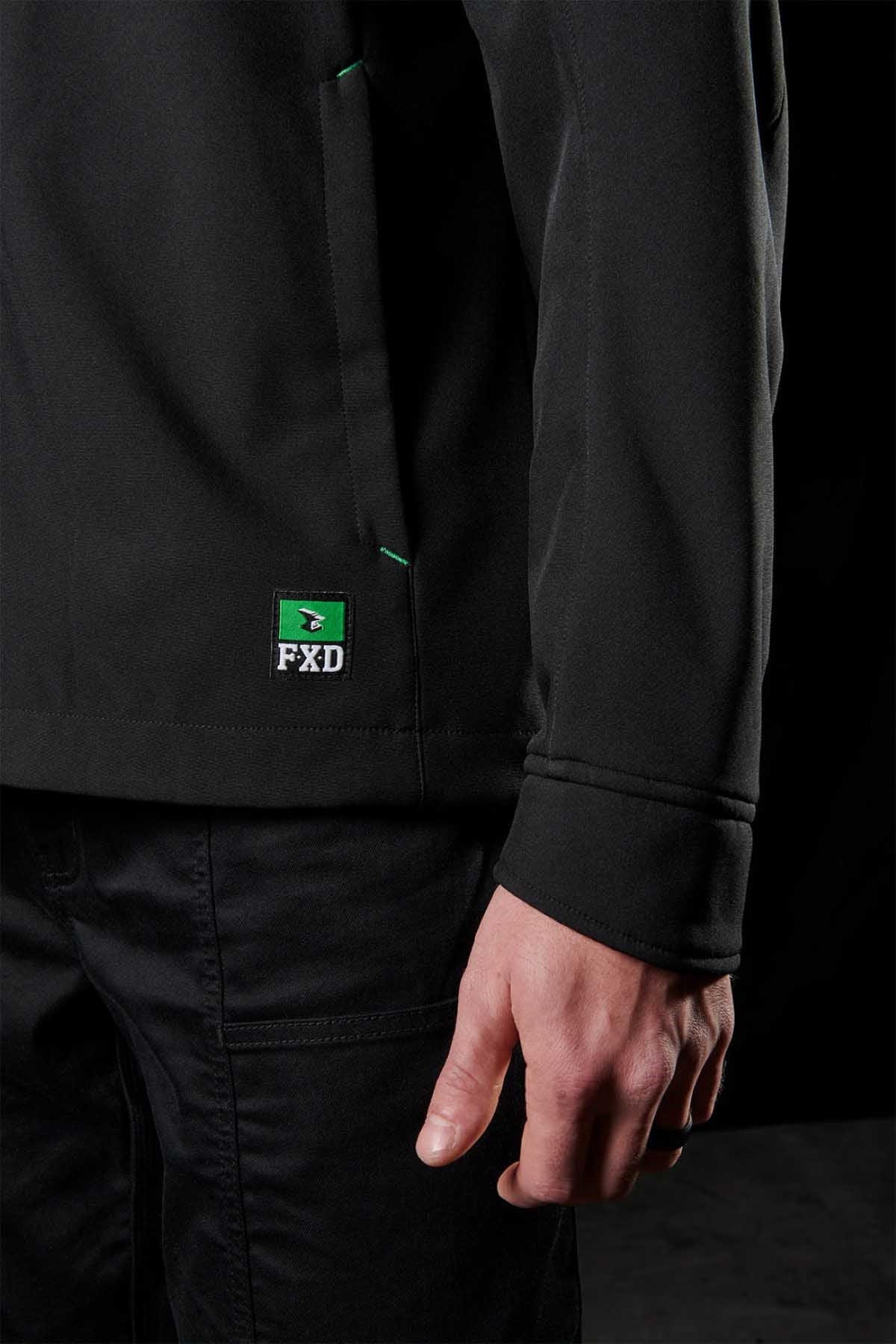 A person clad in a black FXD WO-3 Soft Shell Work Jacket featuring breathable, wind and water-resistant material, paired with matching pants. The photo captures the jacket's side and the person's hand against a dark backdrop. It’s a perfect blend of style with 50+ UPF protection.
