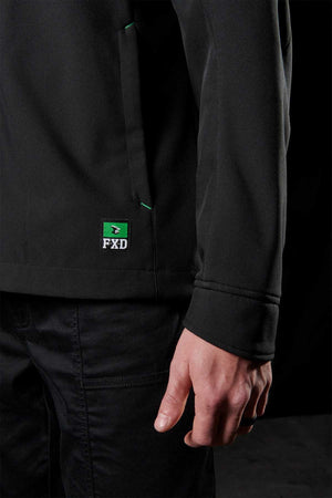 A person clad in a black FXD WO-3 Soft Shell Work Jacket featuring breathable, wind and water-resistant material, paired with matching pants. The photo captures the jacket's side and the person's hand against a dark backdrop. It’s a perfect blend of style with 50+ UPF protection.