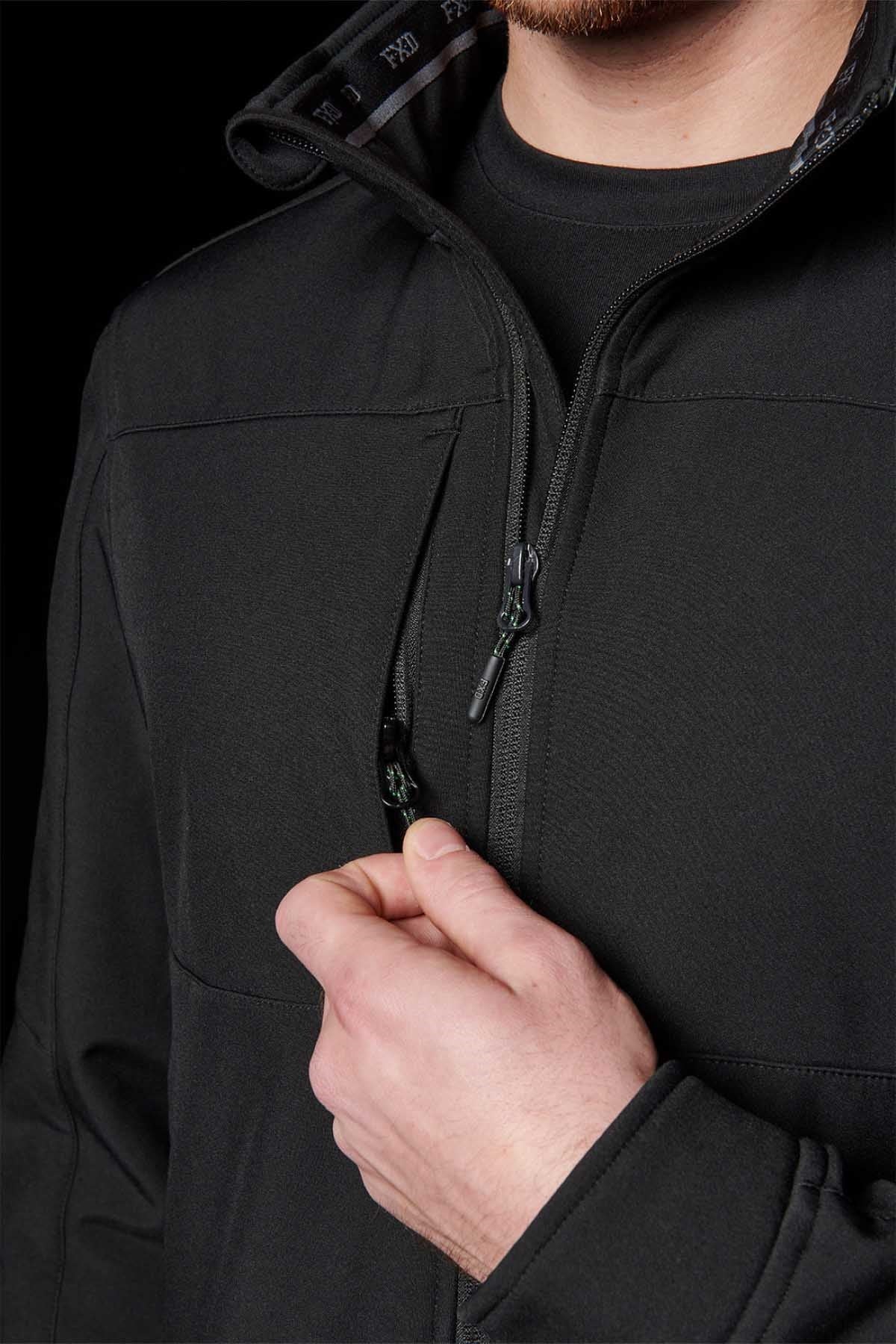 A person in a FXD WO-3 Soft Shell Work Jacket, featuring a high collar and black texture, partially unzips it. Against the black background, only their lower face is visible. This jacket is breathable, wind and water-resistant, and provides 50+ UPF protection.