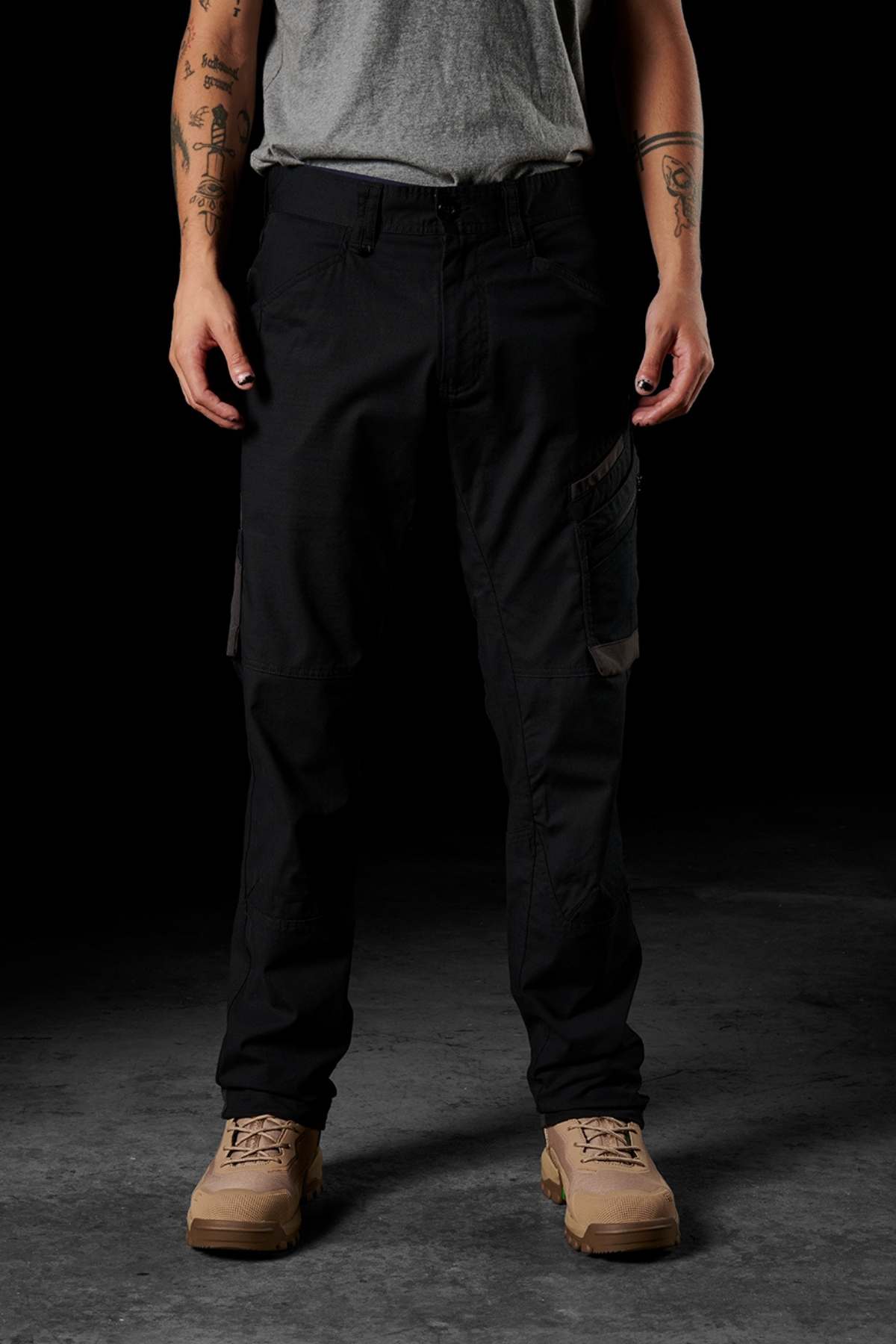 A person stands against a dark background, wearing a gray t-shirt, FXD WP-10 work pants with multi-functional cargo pockets, and tan combat boots. Their arms are tattooed, with their hands resting by their sides.
