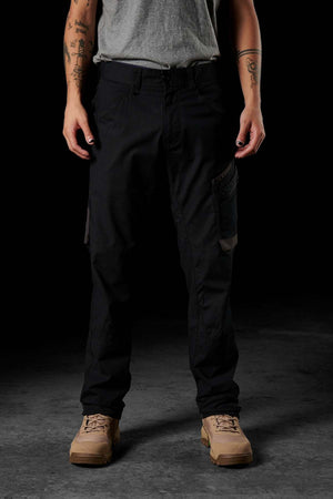 A person stands against a dark background, wearing a gray t-shirt, FXD WP-10 work pants with multi-functional cargo pockets, and tan combat boots. Their arms are tattooed, with their hands resting by their sides.