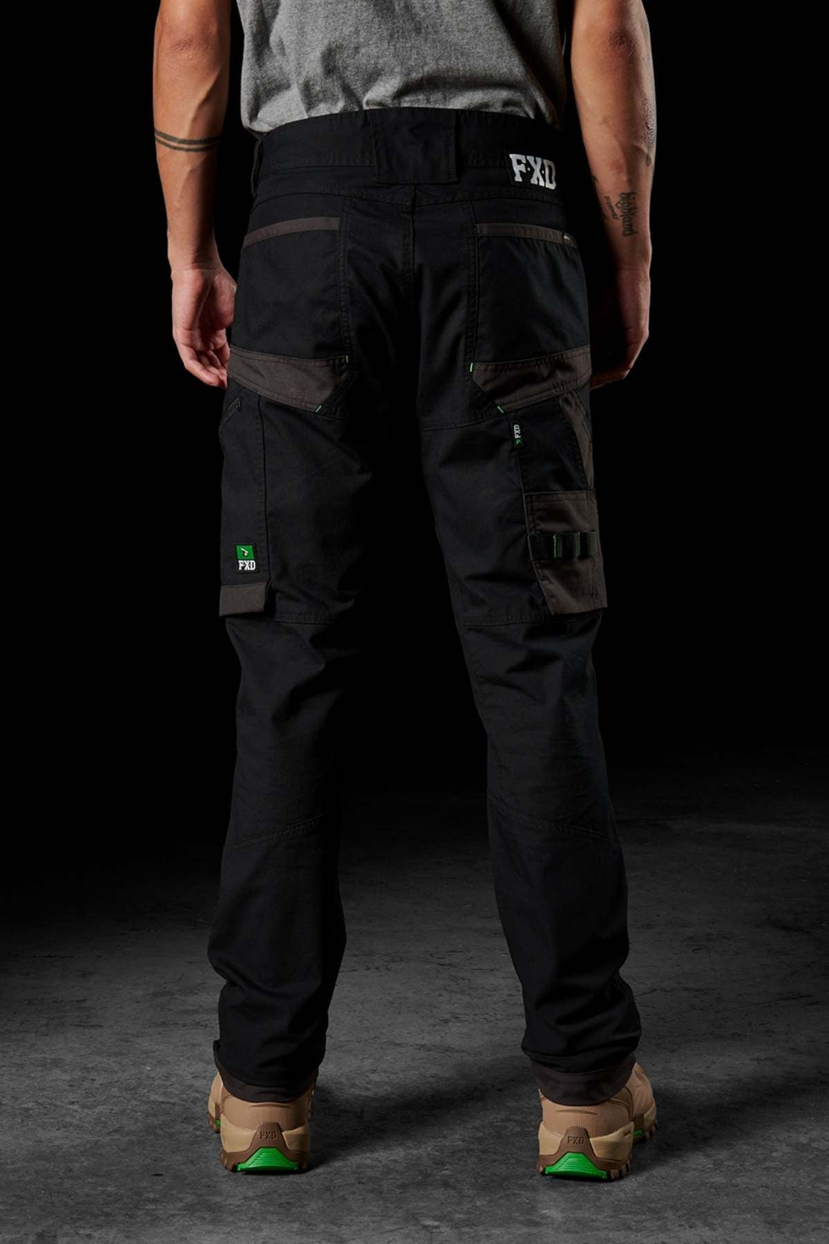A person wearing FXD WP-10 pants, featuring multi-functional cargo pockets, and a gray shirt stands with their back facing the camera. The individual also sports brown and green shoes, along with visible tattoos on their arms. The dark background accentuates the person's appearance.