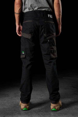 A person wearing FXD WP-10 pants, featuring multi-functional cargo pockets, and a gray shirt stands with their back facing the camera. The individual also sports brown and green shoes, along with visible tattoos on their arms. The dark background accentuates the person's appearance.