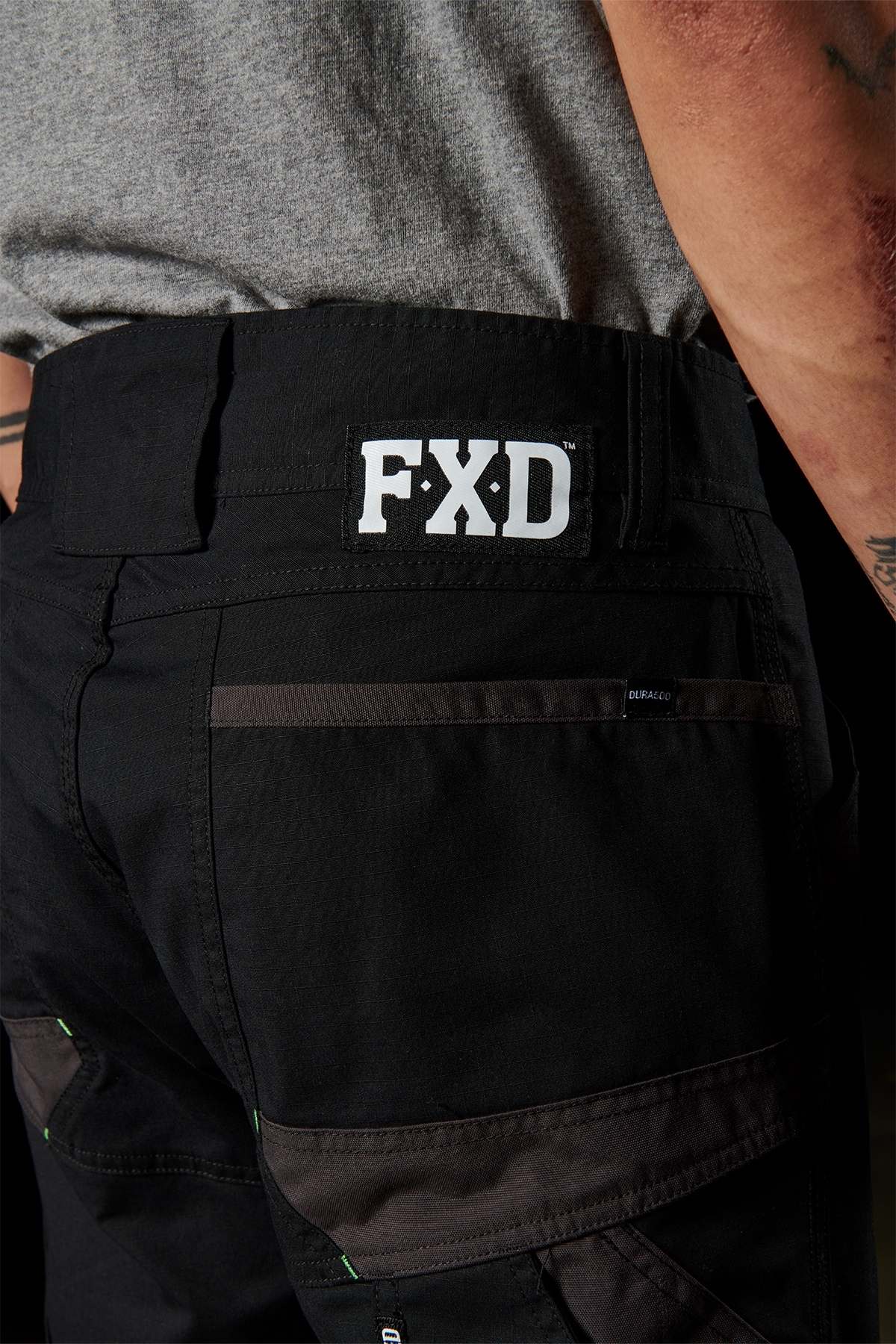 Close-up of an individual wearing the FXD WP-10 pants, showcasing a prominent "FXD" logo on the waistband. The black pants, made from 2-way stretch fabric, are paired with a grey t-shirt and highlight multi-functional cargo pockets along with reinforced stitching. The person also has a tattoo on their forearm.