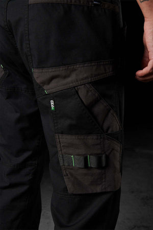 A close-up shot features a person's leg adorned in FXD WP-10 pants, showcasing black, multi-functional cargo pockets with adjustable straps and green threading. The 2-way stretch fabric provides enhanced comfort. The individual stands against a dark background, with their hand partially visible on the right side of the image.