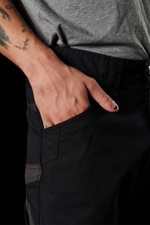 Close-up of a person wearing FXD WP-10 pants in black, with one hand in the multi-functional cargo pocket. The individual sports a tattoo on their forearm and has a black-painted fingernail. They are also dressed in a gray shirt against a predominantly black background.