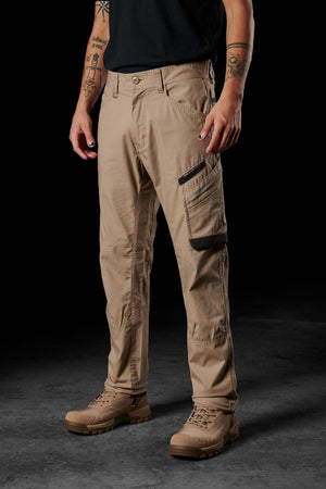 A person stands against a dark background wearing beige FXD WP-10 pants featuring multi-functional cargo pockets and light brown hiking boots. Tattoos are visible on their arms, and they are dressed in a black shirt, though the upper body is partially cropped out of the frame.