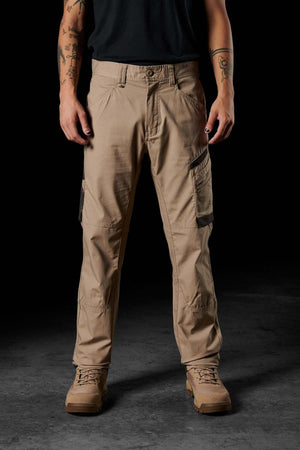 A person wearing FXD WP-10 pants, a dark green shirt, and tan boots stands against a dark background. Only their lower body and arms are visible, adorned with various tattoo designs. The FXD WP-10 pants showcase multi-functional cargo pockets, contributing practicality to their rugged style.