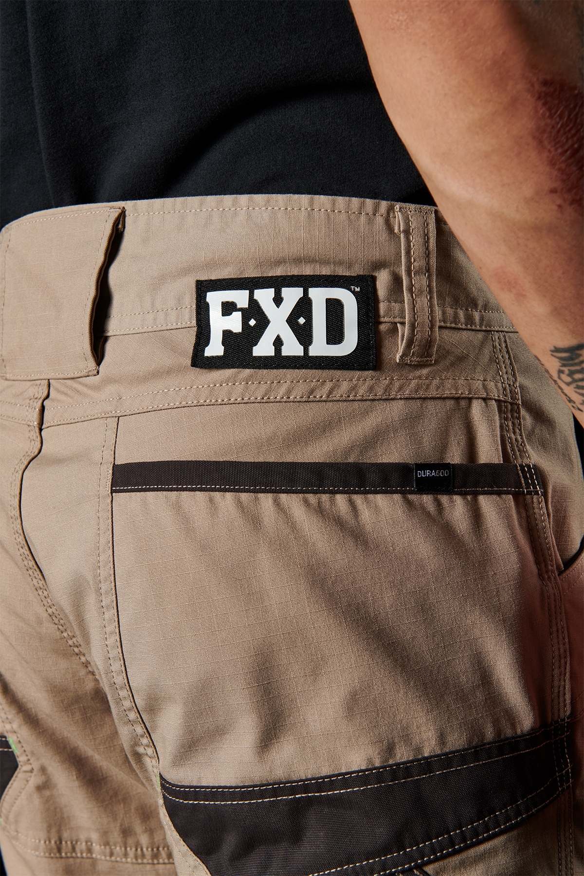 Close-up of a person wearing the FXD WP-10 work pants in beige and black, prominently displaying the "FXD" brand logo on the back. These versatile pants feature a utility pocket, multi-functional cargo pockets, and 2-way stretch fabric. The person is seen from the waist down, paired with a black shirt.