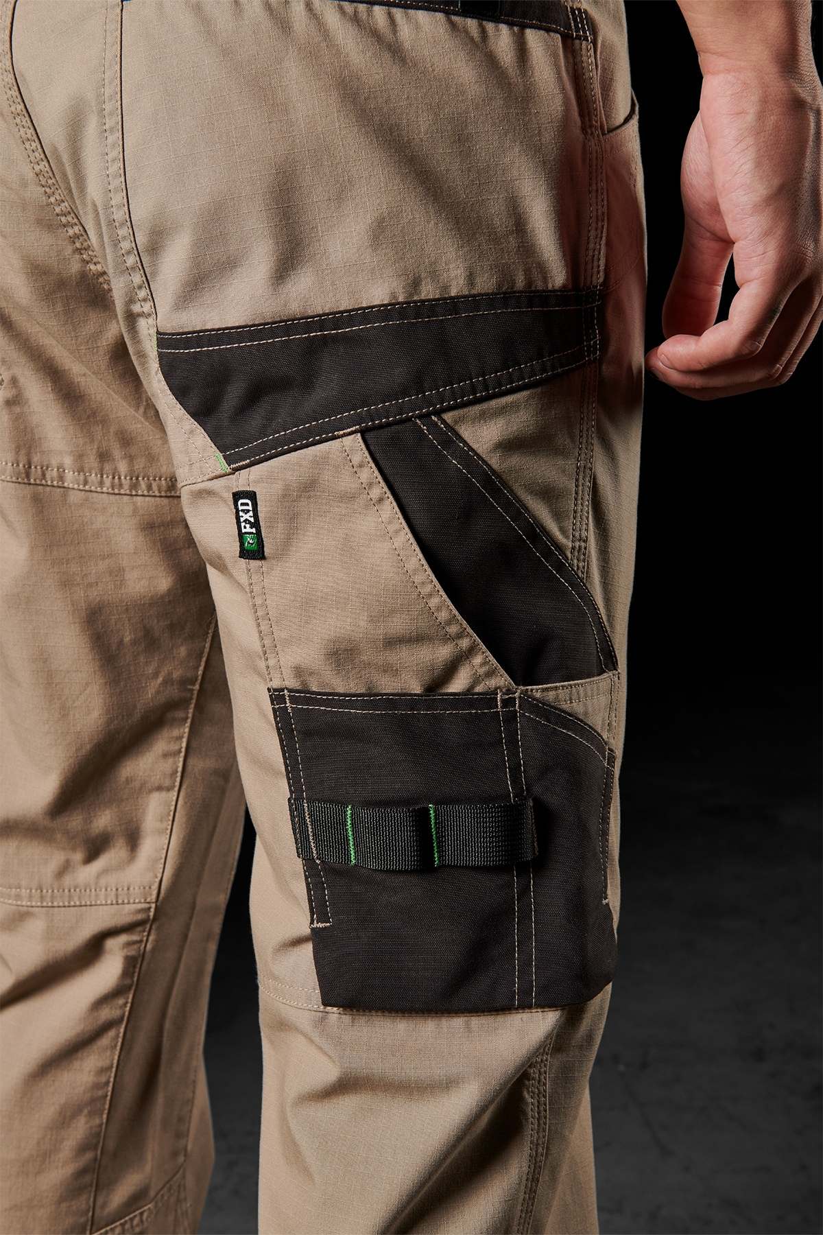 A close-up captures a person showcasing the FXD WP-10 pants, which feature distinctive black accents and versatile cargo pockets. The 2-way stretch fabric and reinforced stitching are prominently highlighted as the individual's right hand grazes the upper pocket. The dark background enhances the focus on the durability and design of these work pants.
