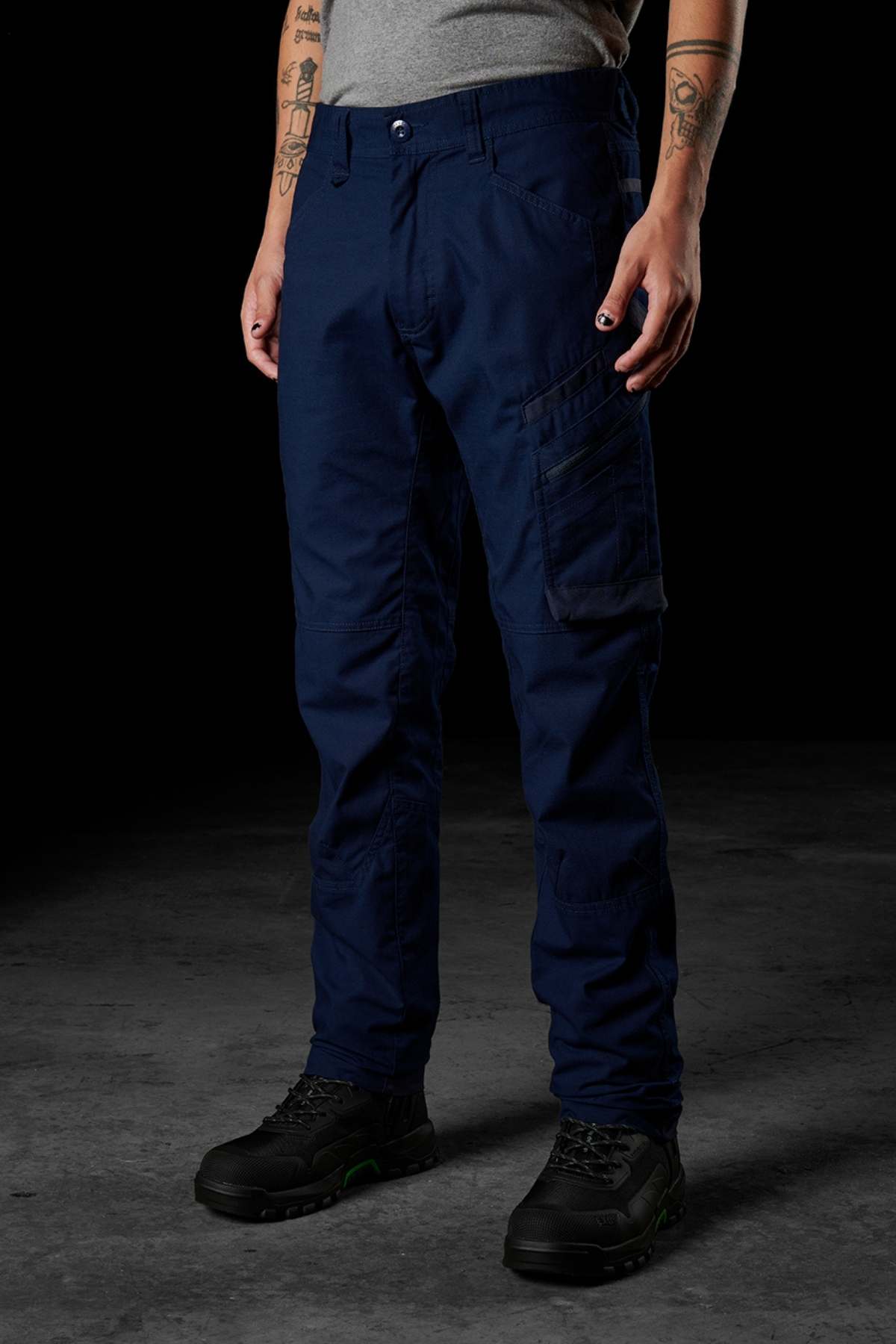 A person dressed in FXD WP-10 dark blue pants featuring multi-functional cargo pockets and a gray shirt stands against a dark background. They have tattoos on their arms, are wearing black and green shoes, and have their nails painted black. The image only shows their lower body as their upper body and head are out of frame.