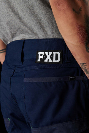 Close-up of a person wearing blue FXD WP-10 work pants featuring multi-functional cargo pockets and a visible logo patch on the waistband. The person, who has tattoos on their forearm, is also dressed in a gray shirt. The background is dark.