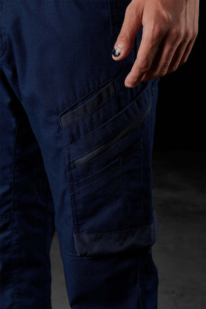 Close-up of a person wearing dark blue FXD WP-10 work pants, featuring multi-functional cargo pockets. The person's hand, adorned with black nail polish, is partially visible resting next to one of the pockets. The background is dark and blurred.