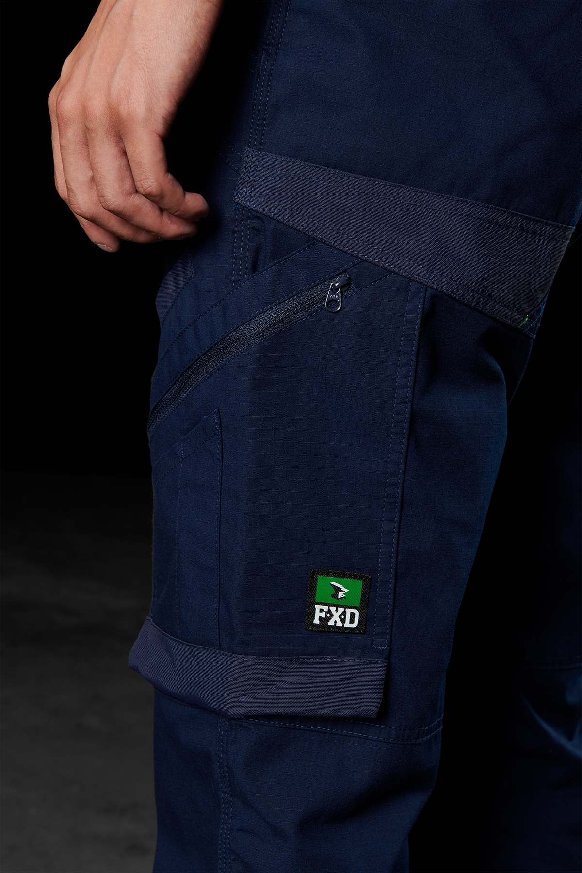 Close-up of a person wearing FXD WP-10 pants in dark blue, featuring detailed stitching and multi-functional cargo pockets, including one with a zipper. A small patch displaying the FXD logo in green, black, and white is visible on the lower part of the pocket. The person's hand rests near the top pocket.