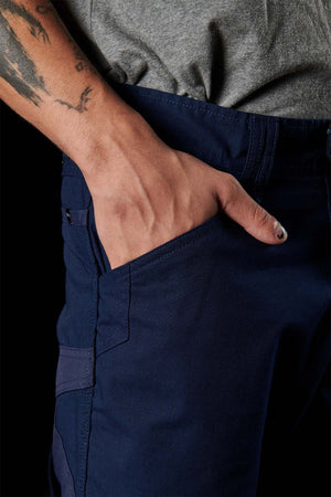 A person is wearing navy blue FXD WP-10 pants with their right hand in the multi-functional cargo pocket, showcasing a tattooed forearm against a dark background. The neatly stitched 2-way stretch fabric pants, featuring side pockets, are paired with a gray shirt. One fingernail of the person is painted black.