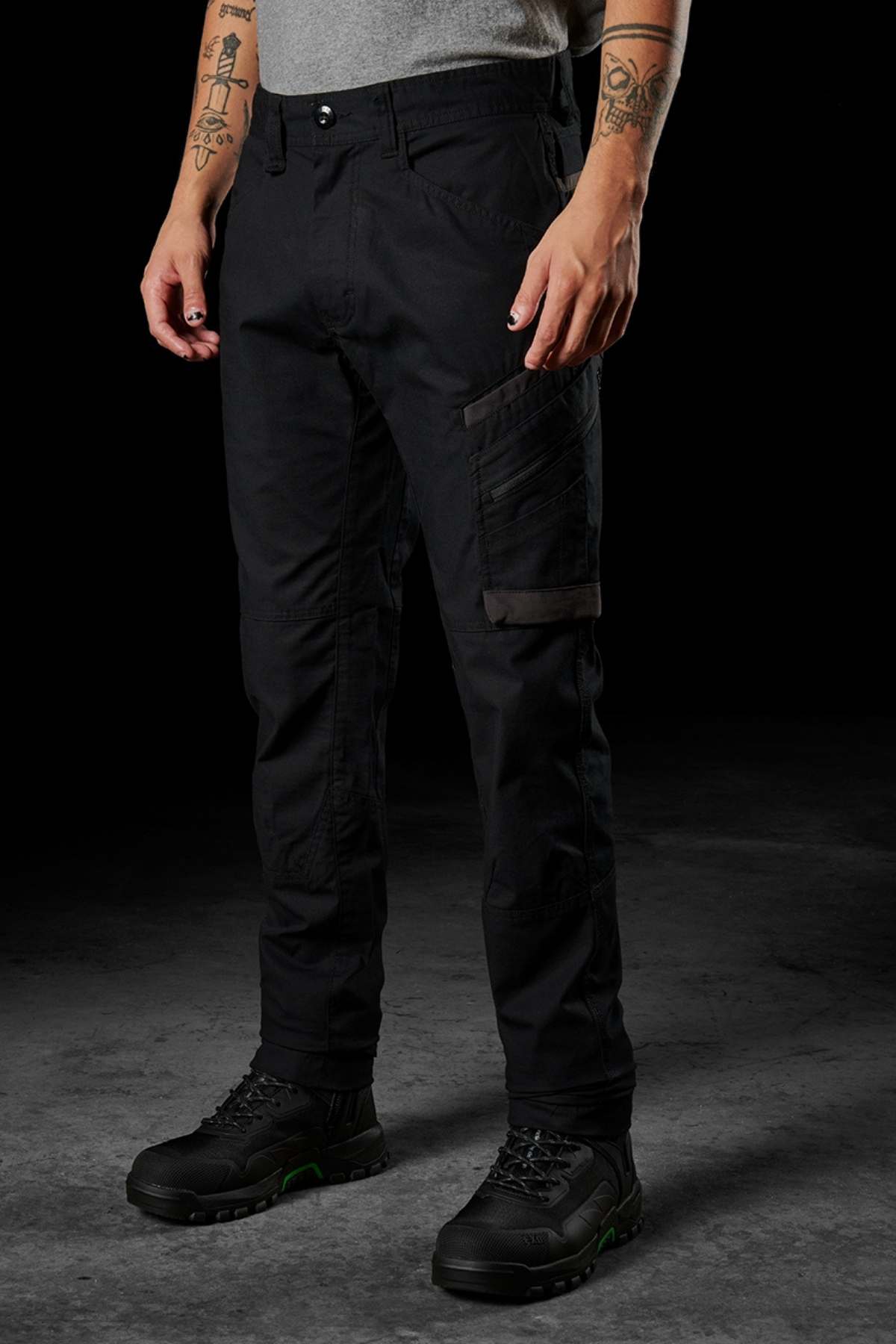 A person stands wearing FXD WP-11 pants in black and black shoes with green accents. Their tattoo-covered arms hang relaxed at their sides. The multi-functional cargo pockets and 2-way stretch fabric of the FXD WP-11 pants blend style and utility strikingly against the dark background.