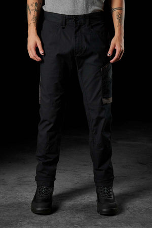 A person is standing against a dark backdrop, dressed in black FXD WP-11 pants featuring multi-functional cargo pockets, black boots, and a grey t-shirt. Tattooed arms are on display, with nails painted black. Their face remains out of view."""