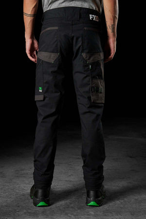 A person wearing the FXD WP-11 pants, which feature multi-functional cargo pockets and green accents, stands with their back to the camera. They are also sporting black shoes with green soles. The backdrop is a dark, neutral surface.
