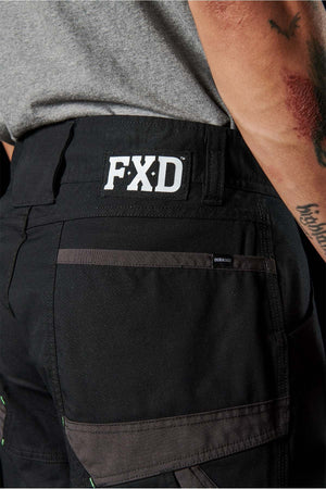 Close-up of a person's back wearing black FXD WP-11 pants and a light gray shirt. The FXD WP-11 pants showcase a bold white FXD logo on the waistband, multi-functional cargo pockets, and distinctive green stitching. Crafted with 2-way stretch fabric for enhanced comfort, the person also sports a tattoo on their left arm.