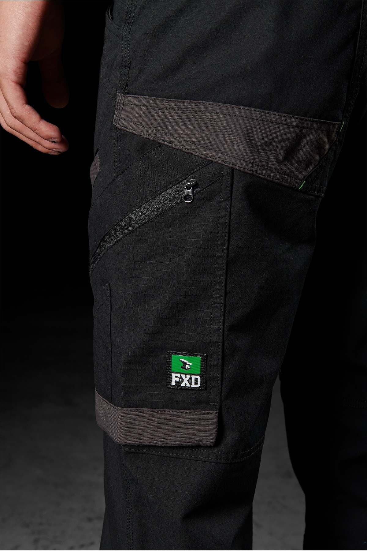 A close-up of a person wearing FXD WP-11 pants shows black work pants equipped with multi-functional cargo pockets and multiple zippers. The pants feature a small green and black logo patch with "FXD" on the side pocket. The background is dark, revealing only a portion of the person's hand.