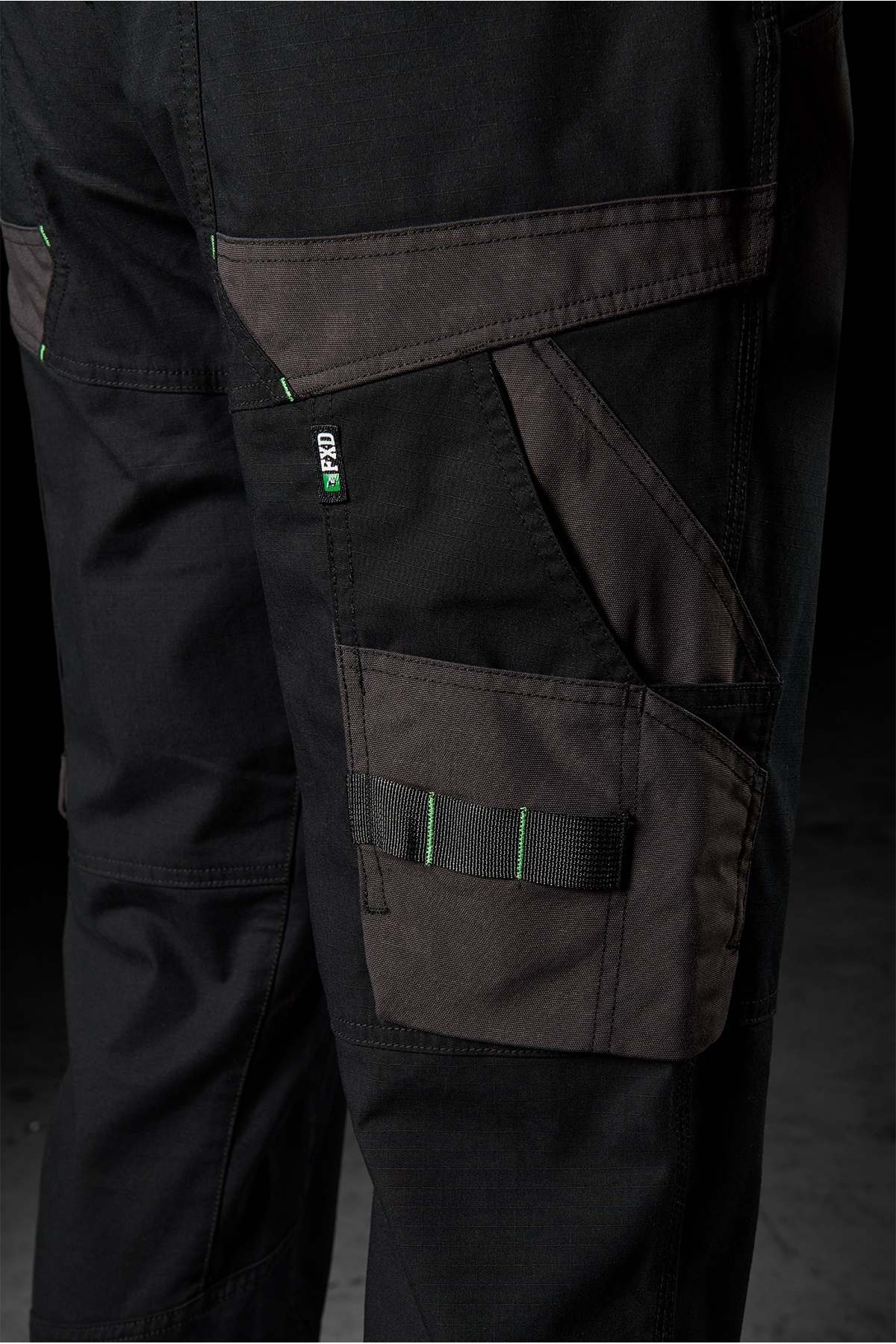 Close-up of FXD WP-11 work pants in black with dark grey accents, showcasing multi-functional cargo pockets and a green and white "PRO" label. These pants feature a robust design with 2-way stretch fabric ideal for heavy-duty tasks, reinforced stitching, and practical storage solutions. The background is dark.