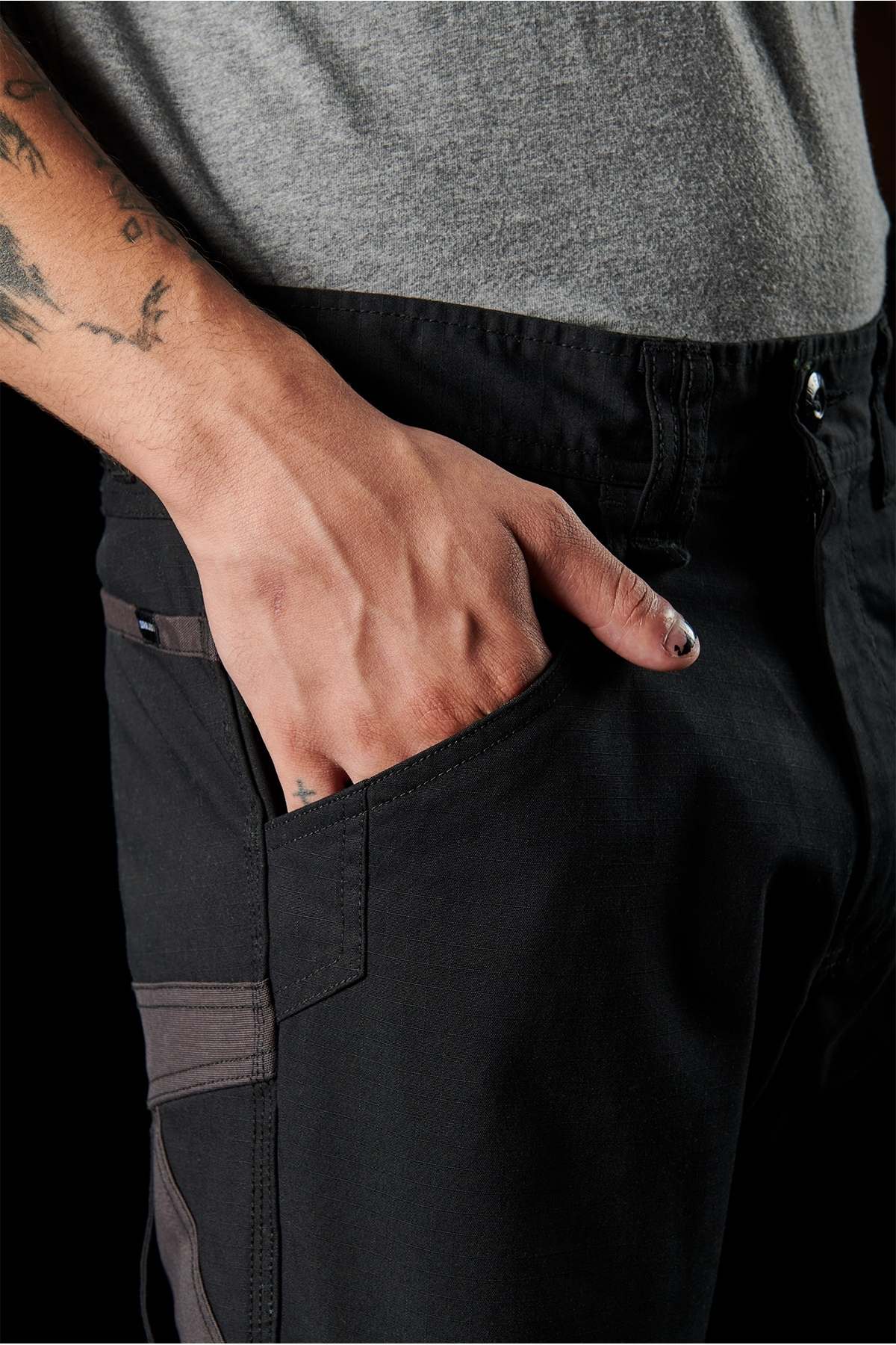 A close-up shot of a tattooed hand resting in the pocket of FXD WP-11 pants highlights the multi-functional cargo pockets. The person, sporting black-painted nails, wears a gray shirt against a black background.